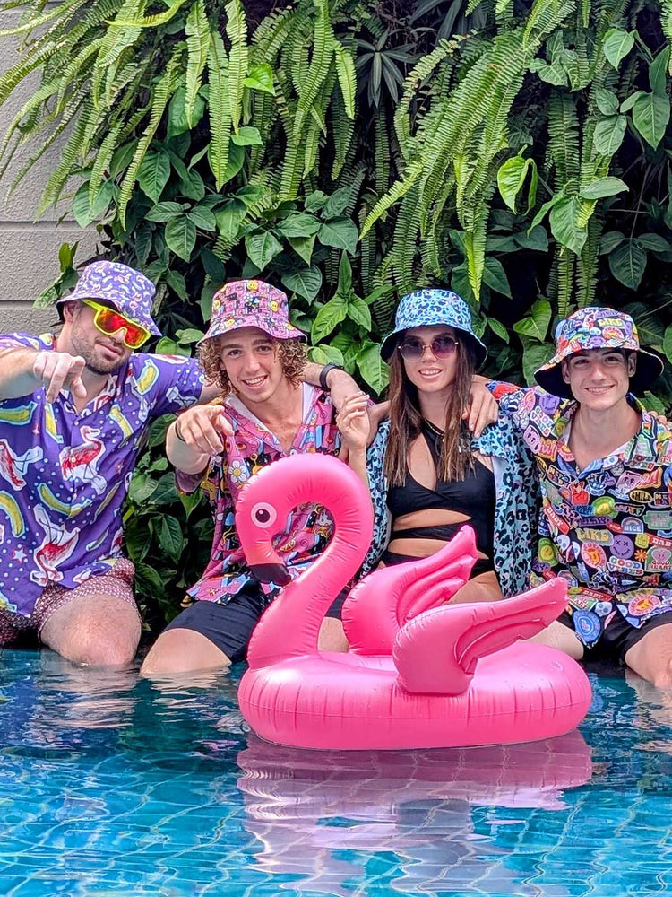 Four young adults in colourful, shit shirt outfits and bucket hats sit at the edge of a pool with a pink inflatable flamingo. Lush green plants are in the background, and everyone is smiling and having fun.