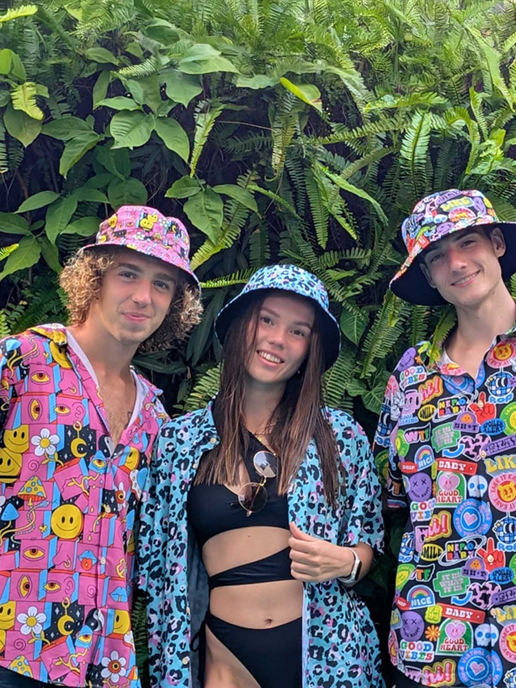 Three young adults pose in front of lush green ferns. Two men wear colourful bucket hats and matching patterned shit shirts, while the woman in the centre wears a black swimsuit with a blue animal print shit shirt and a matching bucket hat. All are smiling.