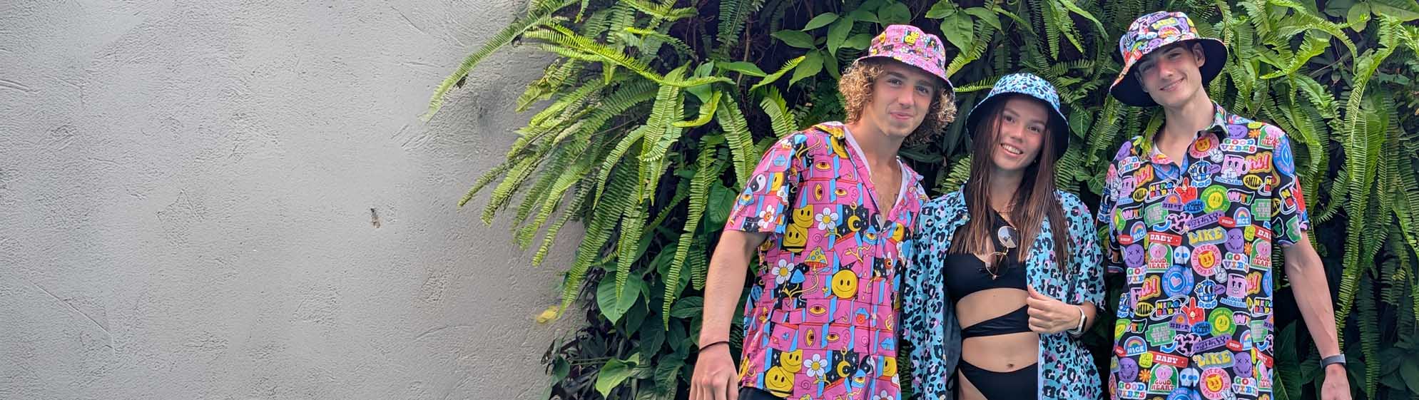 Three people stand in front of a leafy green wall, wearing colourful matching bucket hats and shit shirts with playful patterns. The woman in the centre wears a black bikini top and open shit shirt. All are smiling at the camera.