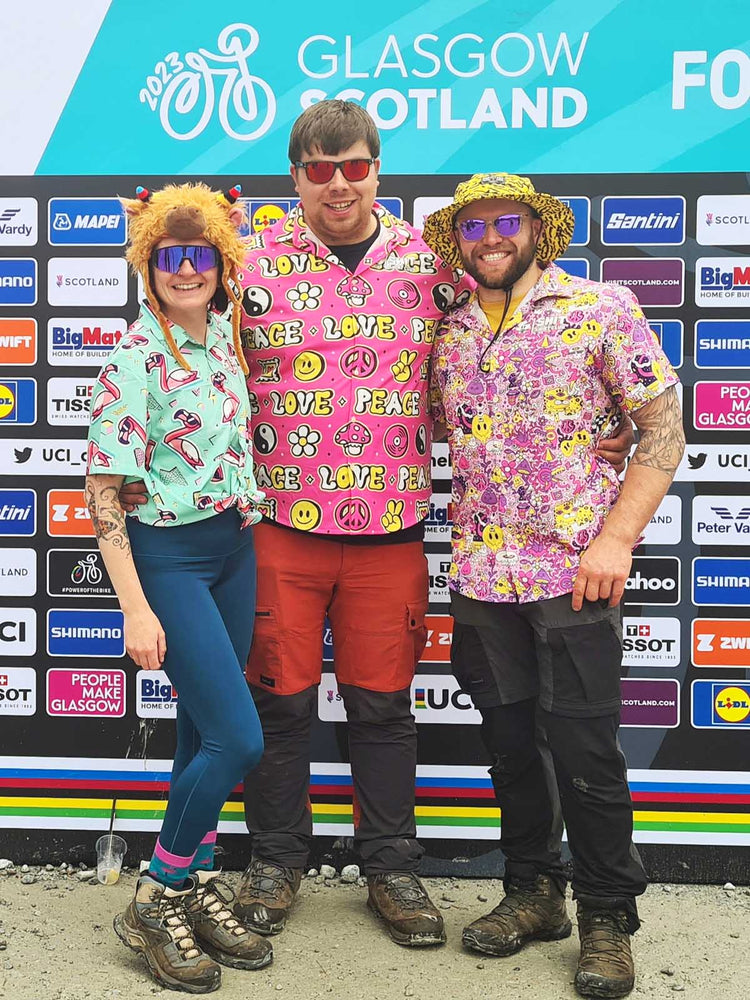 Three people in colourful, playful shit shirts and hats stand smiling in front of a backdrop with sponsor logos at the 2023 UCI Cycling World Championships in Glasgow, Scotland.